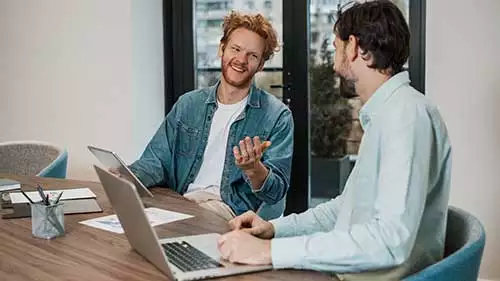 Två män vid ett skrivbord talar med varandra. Den ena sitter vid en laptop.